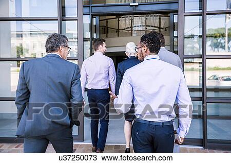 Stock Image - Rear view of businessmen and woman arriving at office building. Fotosearch
