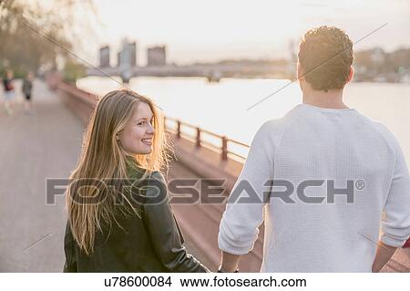 Picture - Rear view of couple strolling on waterfront at sunset, Battersea Park, London, UK. Fotosearch