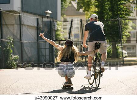 Stock Photo - Rear view of female skateboarder and male cyclist on basketball court. Fotosearch