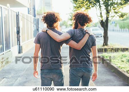 Stock Image - Rear view of identical male adult twins strolling on sidewalk. Fotosearch