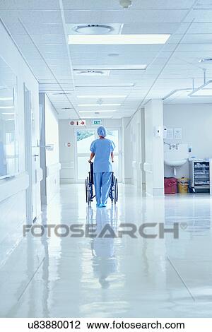 Stock Image - Rear view of nurse pushing wheelchair along hospital corridor. Fotosearch
