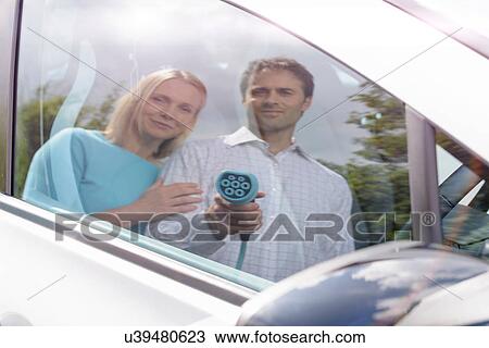 Stock Image - Reflection of man and woman with electric, car holding charging plug. Fotosearch