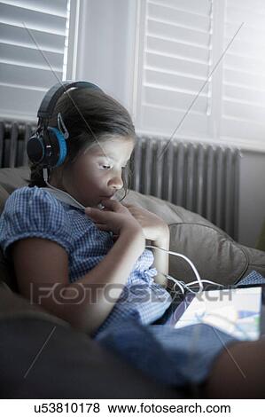 Schoolgirl sitting on sofa using digital tablet and listening to headphones View Large Photo Image Stock Photo - Schoolgirl sitting on sofa using digital tablet and listening to headphones. Fotosearch