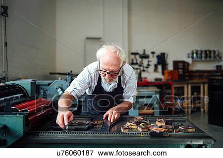Stock Photo - Senior craftsman bending over and using wooden letters and letterpress machine in book arts workshop. Fotosearch