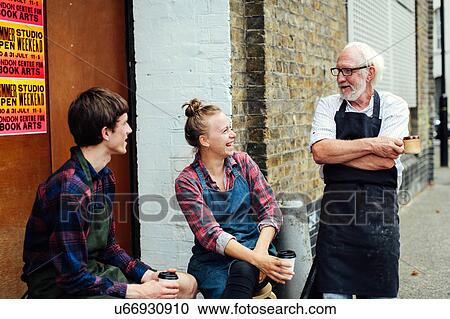 Stock Image - Senior craftsman chatting and laughing with young craftswoman and craftsman outside print workshop. Fotosearch