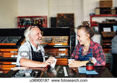 Stock Photo - Senior craftsman chatting and laughing with young craftswoman in book arts workshop. Fotosearch