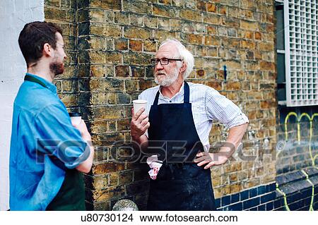 Senior craftsman drinking coffee and chatting to young man outside workshop View Large Photo Image Picture - Senior craftsman drinking coffee and chatting to young man outside workshop. Fotosearch