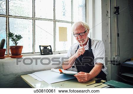 Stock Image - Senior craftsman holding pencil and looking at clipboard in print workshop. Fotosearch