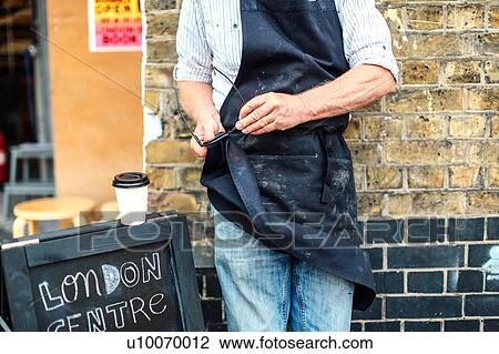 Stock Image - Senior craftsman outside workshop, cleaning glasses with his apron, mid section. Fotosearch