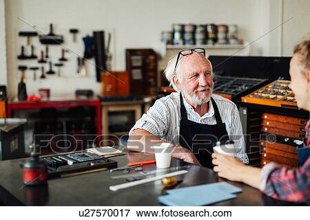 Stock Photo - Senior craftsman sitting at table with young craftswoman and smiling in book arts workshop. Fotosearch
