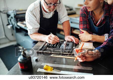 Stock Photograph - Senior craftsman using letterpress equipment and teaching young craftsman and craftswoman in print workshop. Fotosearch