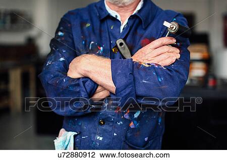 Stock Image - Senior craftsman with arms folded in print workshop, holding ink roller and wearing paint-splattered overalls, mid section. Fotosearch