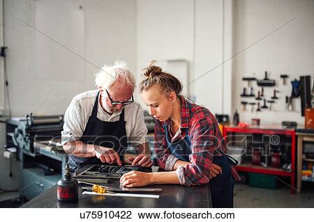 Stock Image - Senior craftsman working on letterpress with young craftswoman in print workshop. Fotosearch