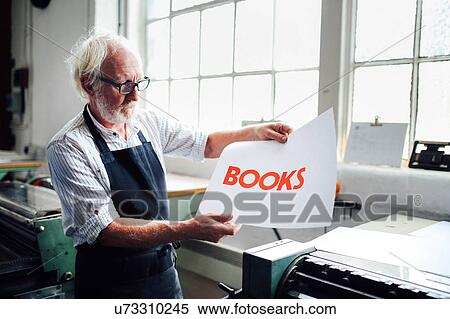 Stock Photography - Senior craftsman/technician holding up letterpress print in book arts workshop. Fotosearch