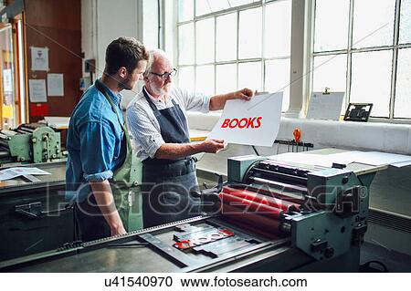 Stock Image - Senior craftsman/technician showing young man letterpress print in book arts workshop. Fotosearch