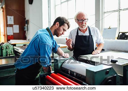 Stock Image - Senior craftsman/technician supervising young man on letterpress machine in book arts workshop. Fotosearch