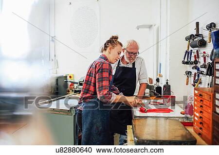 Stock Image - Senior craftsman/technician supervising young woman as she uses ink roller for letterpress printing in book arts workshop. Fotosearch