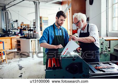 Stock Photograph - Senior craftsman/technician supervising young man on letterpress machine in book arts workshop. Fotosearch