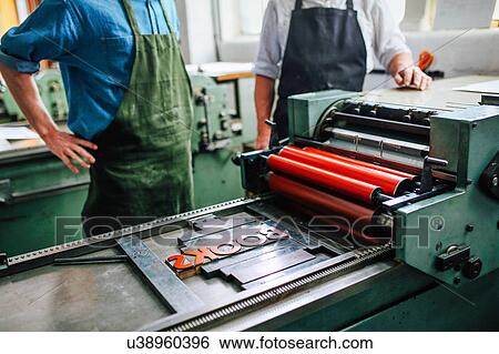 Stock Photograph - Senior craftsman/technician supervising young man on letterpress machine in book arts workshop, mid section. Fotosearch