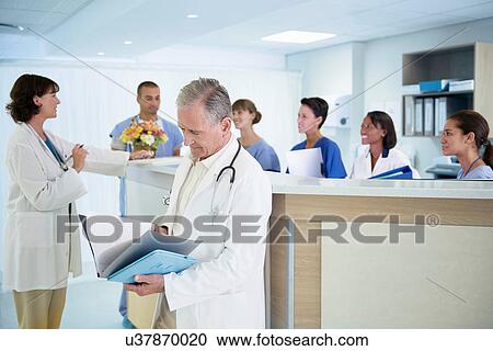 Stock Image - Senior doctor reading medical notes at hospital nurses station. Fotosearch