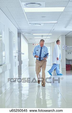 Stock Photography - Senior doctor walking along hospital corridor reading medical notes. Fotosearch