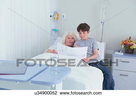 Stock Image - Senior female patient in hospital bed looking at grandson's drawing. Fotosearch
