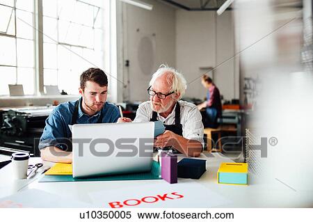 Stock Photography - Senior male craftsman and young man looking at laptop in book arts workshop. Fotosearch