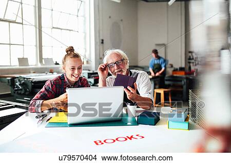 Senior male craftsman laughing and looking at laptop with young woman in book arts workshop View Large Photo Image Picture - Senior male craftsman laughing and looking at laptop with young woman in book arts workshop. Fotosearch