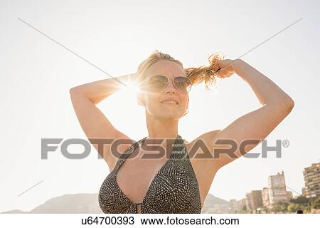 Stock Image - Sunlit portrait of woman tying back hair at beach, Alicante, Spain. Fotosearch