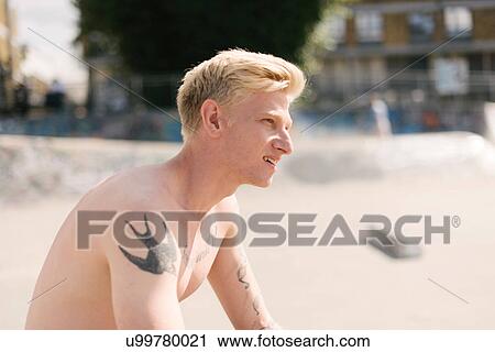 Stock Image - Tattooed young male skateboarder sitting in skatepark. Fotosearch