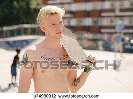 Stock Image - Tattooed young man carrying skateboard on shoulder in skatepark. Fotosearch