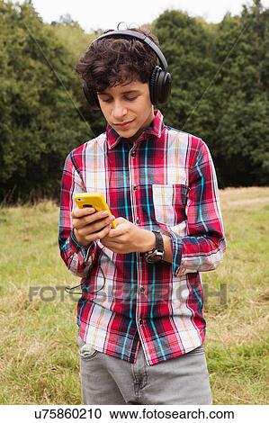 Stock Image - Teenage boy, standing in fields, wearing headphones, using smartphone. Fotosearch