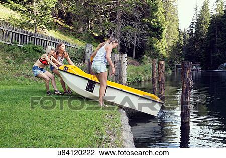Stock Image - Three adult female friends launching rowing boat into lake, Sattelbergalm, Tirol, Austria. Fotosearch