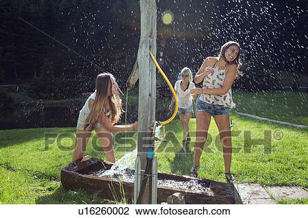 Stock Image - Three adult female friends play fighting with water hose in garden. Fotosearch