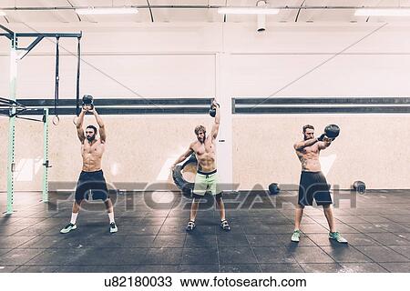 Stock Image - Three young male cross trainers training with kettlebells in gym. Fotosearch
