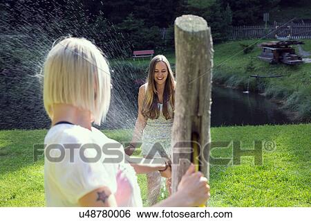 Stock Photograph - Two adult female friends play fighting sprinkling water hose in garden. Fotosearch