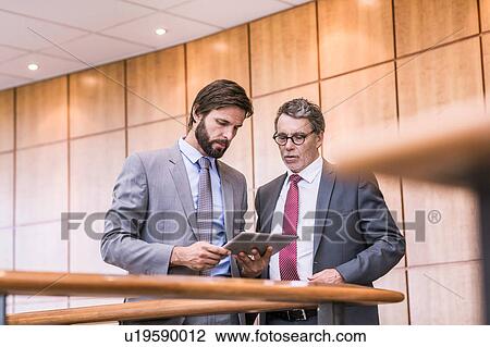 Stock Image - Two businessmen reading digital tablet on office balcony. Fotosearch
