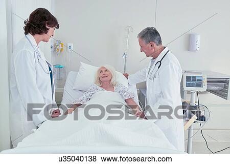 Two doctors talking to senior female patient in hospital bed View Large Photo Image Stock Photo - Two doctors talking to senior female patient in hospital bed. Fotosearch
