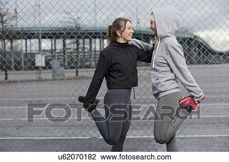 Stock Image - Two female runner friends stretching legs by wire fence. Fotosearch