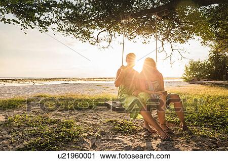 Stock Image - Two women sitting on beach tree swing at sunset, Gili Trawangan, Lombok, Indonesia. Fotosearch