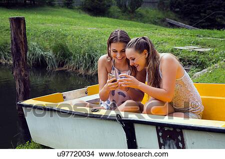 Picture - Two young female friends reading smartphone texts in rowing boat on lakeside. Fotosearch