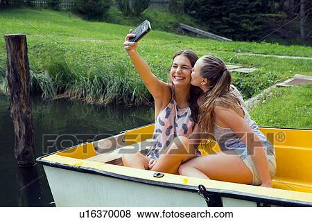 Stock Photo - Two young female friends taking smartphone selfie in rowing boat on lakeside. Fotosearch