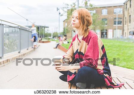 Stock Photo - Woman holding cup sitting cross legged looking away, London, UK. Fotosearch