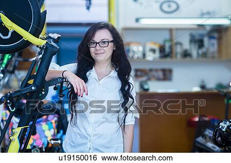 Stock Photograph - Woman in bicycle shop leaning against bike frame looking at camera smiling. Fotosearch