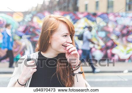 Stock Image - Woman in urban area hand on mouth looking away smiling, London, UK. Fotosearch