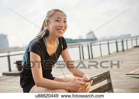 Stock Photo - Woman wearing earphones holding smartphone smiling. Fotosearch