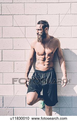Stock Image - Young bare-chested male cross trainer leaning against wall outside gym. Fotosearch
