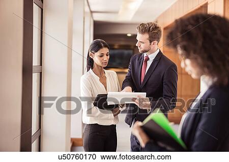 Stock Image - Young businesswoman and businessman reading paperwork in office corridor. Fotosearch
