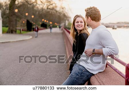 Stock Image - Young couple chatting on waterfront at sunset, Battersea Park, London, UK. Fotosearch