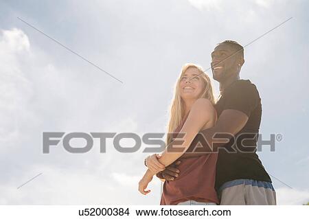 Picture - Young couple with arms around each other against sunlit blue sky. Fotosearch